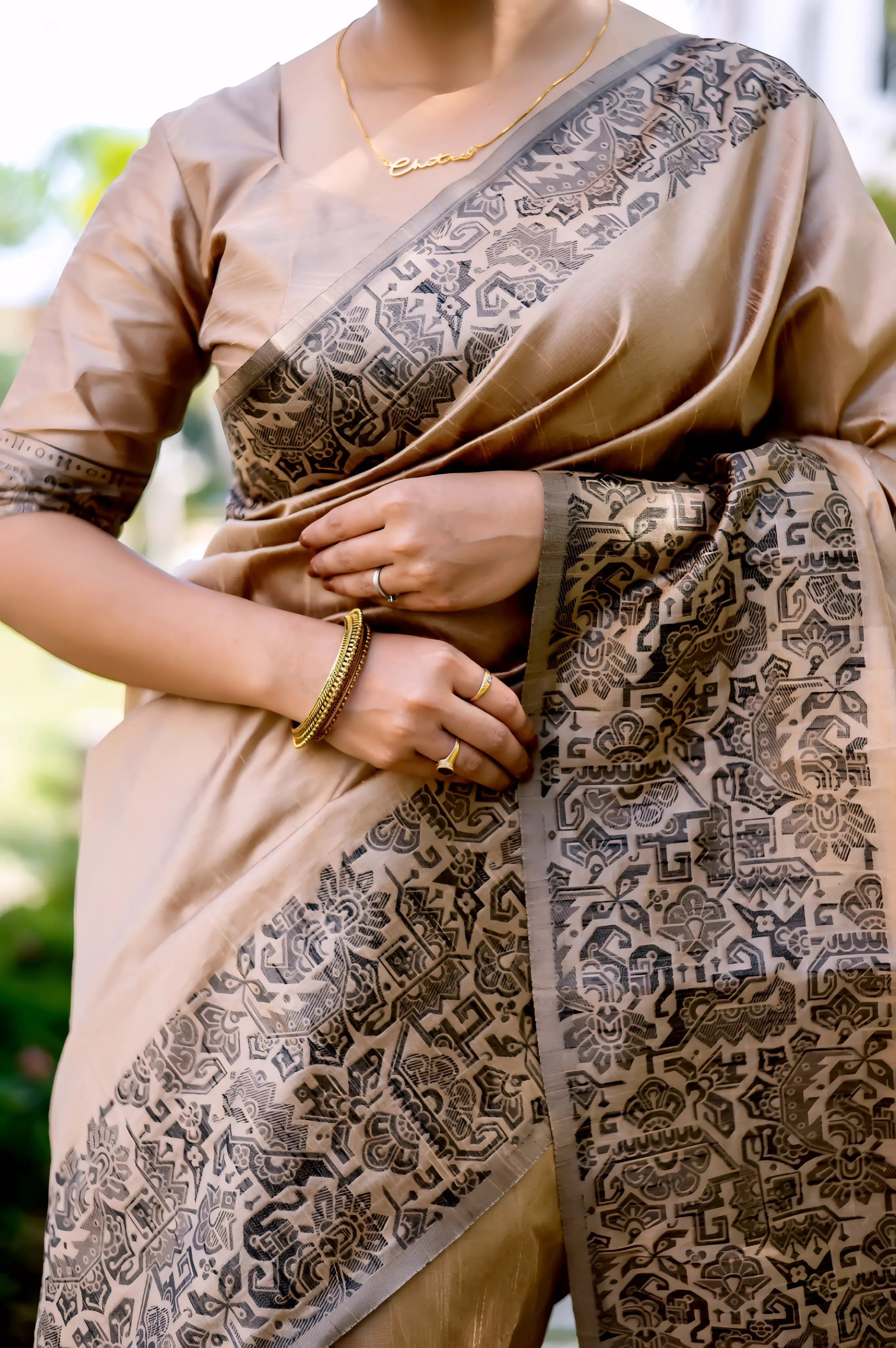 A woman models a luxurious Beige/Taupe Raw Silk Saree. The saree features a wide border and pallu with dense, intricate dark brown/black Zari weaving in a Kalamkari-style floral and paisley pattern. She wears a matching short-sleeve beige blouse and traditional gold jewelry, posing outdoors on a stone pathway.